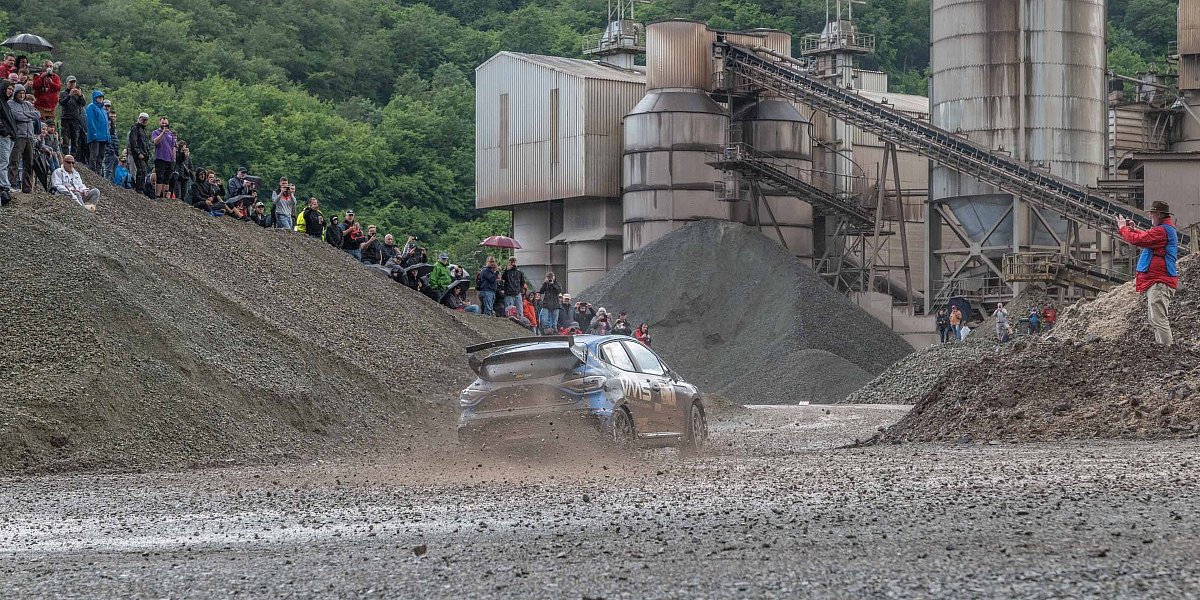 Ein Rallyefahrzeug fährt in einer Schotterkurve auf der Rennstrecke bei der Hunsrück Rallye 2024. Am Rand Zuschauer und ein Fotograf.