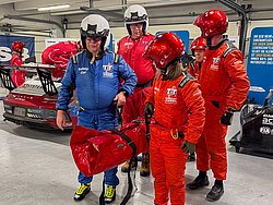 DMSB Medical Days 2026 DMSB Medical Days 2026: Team in roten/blauen Overalls und Helmen trägt eine Tasche, in der eine Person abtransportiert wird; im Hintergrund ein Rennwagen.