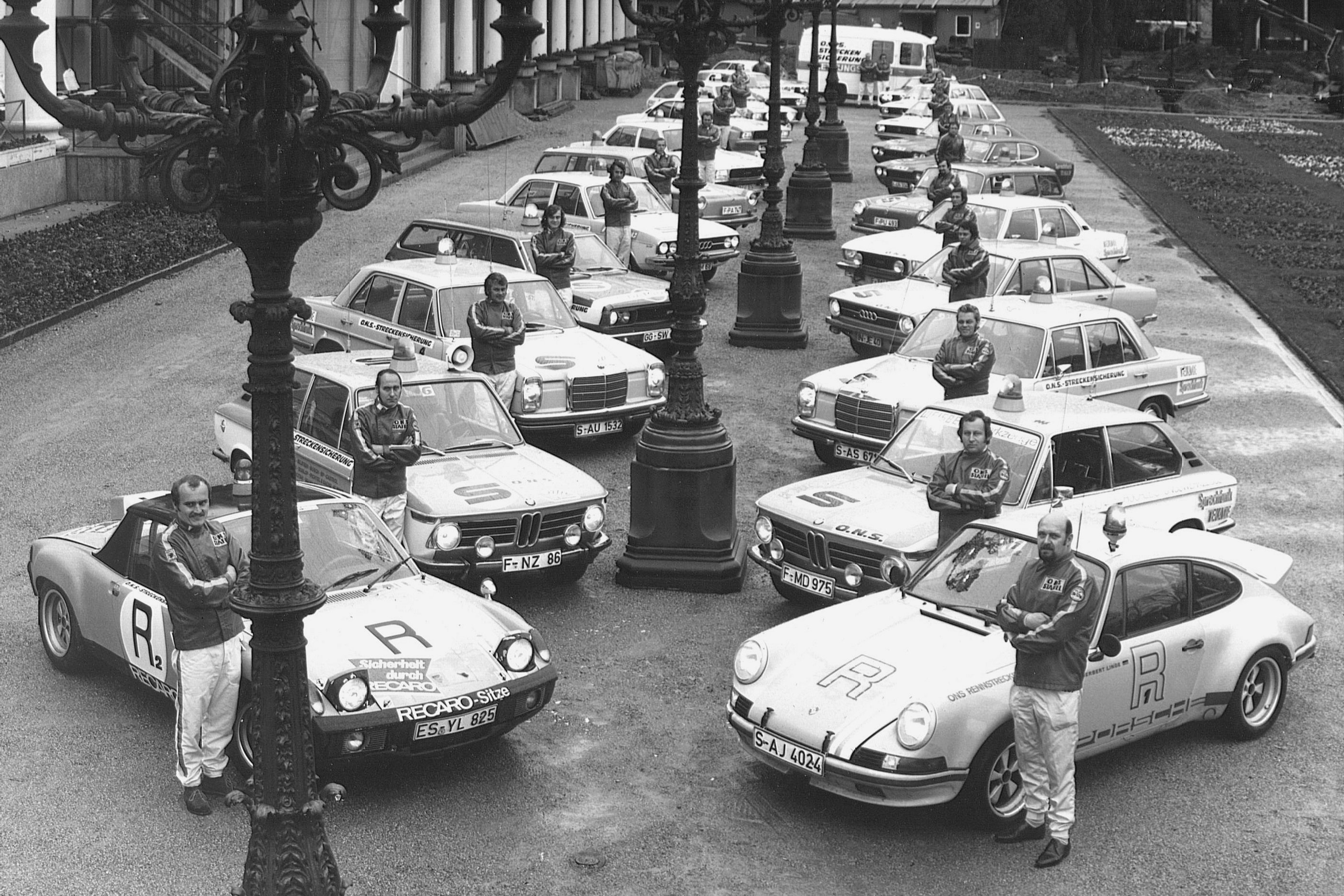 Eine Gruppe von ONS-Staffelaners posiert zum fünfjährigen Jubiläum mit Staffelautos, die in einer Reihe auf einer Straße in der Nähe von verschnörkelten Laternenpfählen stehen.