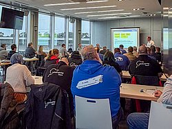 DMSB Medical Days 2026 DMSB Medical Days 2026: Seminarraum mit Gruppe an Tischen; Frontalprojektor mit Präsentation und Redner, Fensterblick auf Rennstrecke und Himmel.