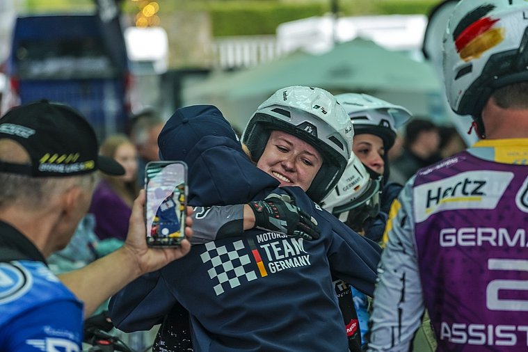 Eine deutsche Trial-Fahrerin im Helm umarmt ein Teammitglied, während ein Fan den emotionalen Moment mit dem Smartphone festhält.