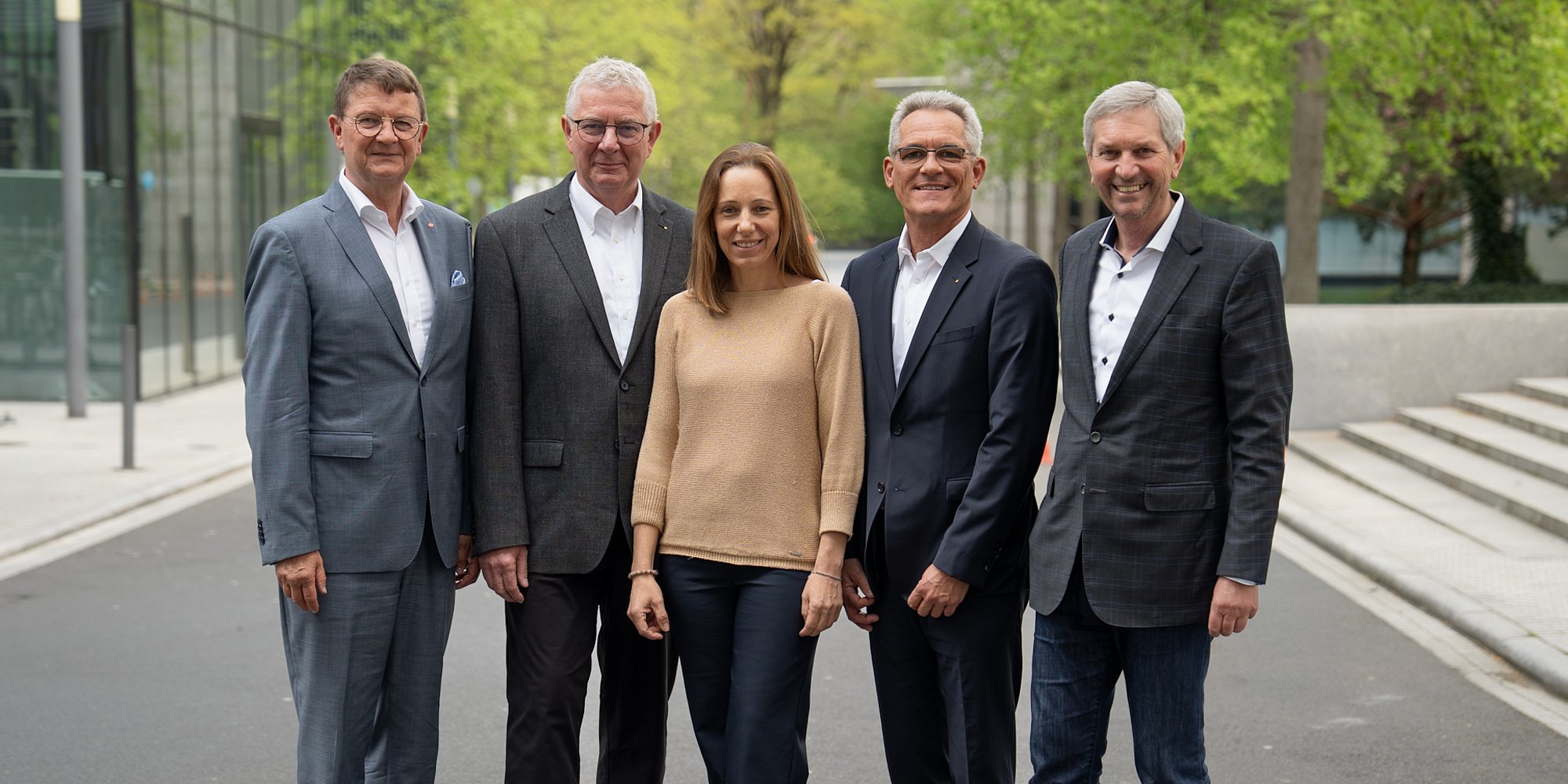 Fünf Personen in Geschäftskleidung stehen im Freien auf einem gepflasterten Weg zusammen und lächeln in die Kamera.