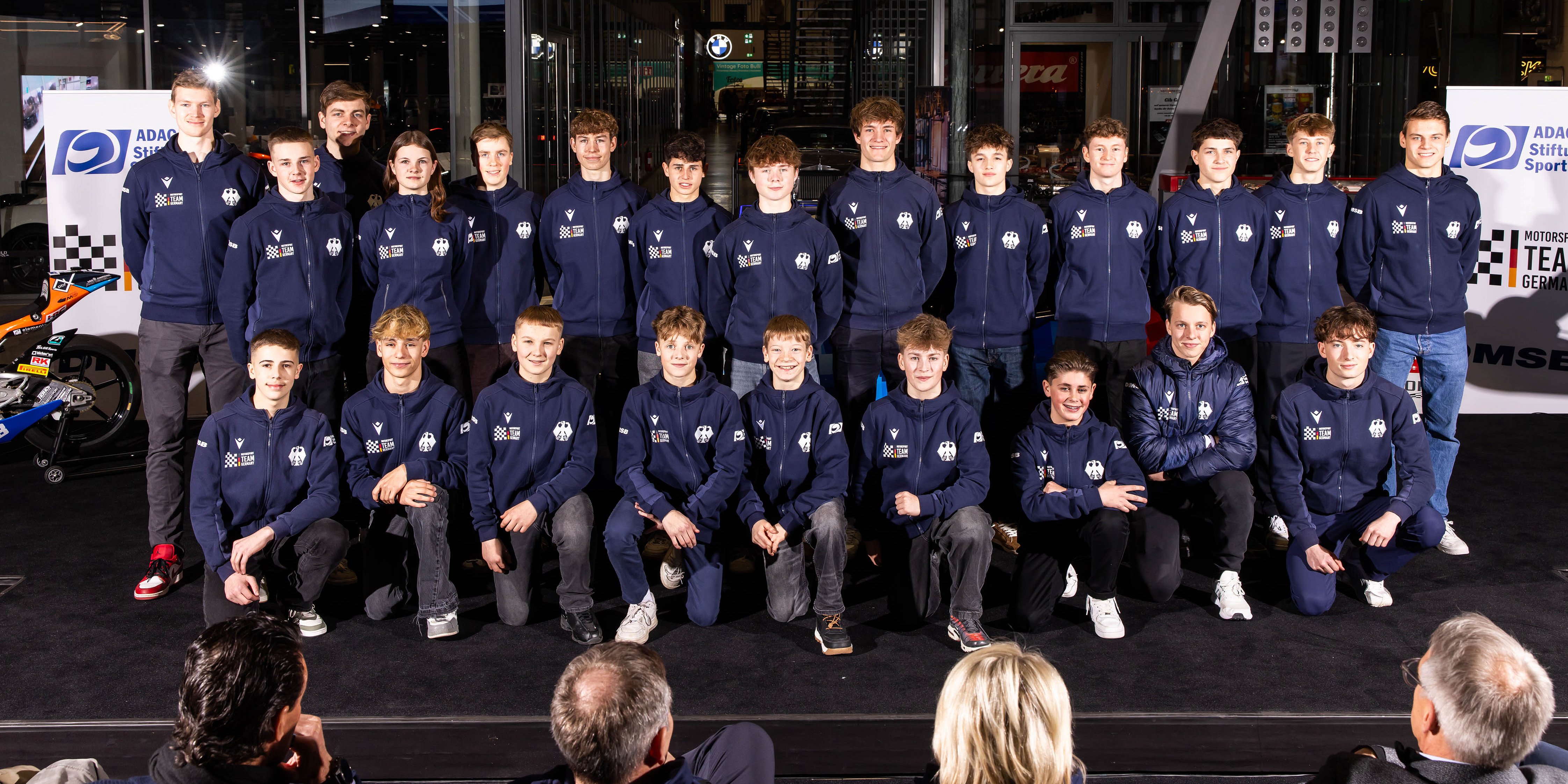 Eine Gruppe von 22 jungen Leuten in passenden blauen Jacken stellt sich auf einer Bühne für ein Gruppenfoto zusammen.
