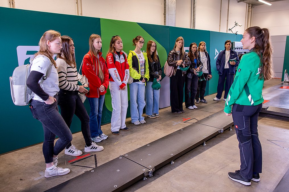 Discover Motorsport – Young Women's Edition 2026 in Oschersleben Die Gruppe steht an der Waage der Technischen Abnahme der DEKRA und hört einer Technischen Kommissarin von DEKRA aufmerksam zu.