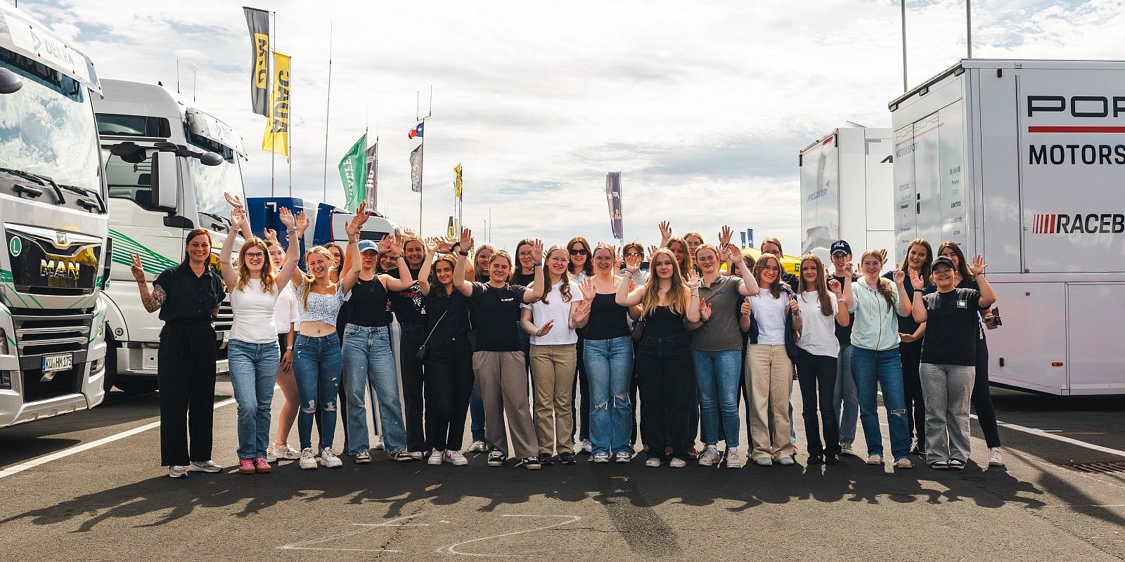 Gruppenfoto von Teilnehmerinnen der Discover Motorsport – Young Women’s Edition 2025 am Nürburgring mit erhobenen Händen vor den DTM-Teamtruckern, dazu das Event-Logo und der Hinweis „Am 23. Juli in Oschersleben – jetzt anmelden“.