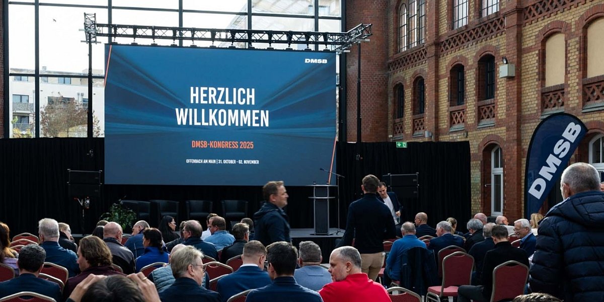 DMSB-Kongress 2025, Eröffnung Podium mit Rednerpult und Leinwand, davor Menschen in Sitzreihen, im Hintergrund Glasscheiben mit Sicht nach draußen. Auf der Leinwand steht: Herzlich Willkommen. DMSB-Kongress 2025. Offenbach am Main | 31. Oktober - 02. November 2025.