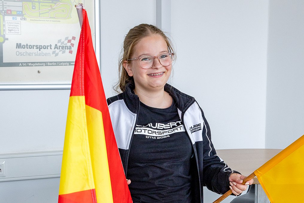 Discover Motorsport – Young Women's Edition 2026 in Oschersleben Eine Teilnehmerin hält bei den Sportwarten der Streckensicherung eine rot-gelbe Flagge und eine gelbe Flagge und lächelt in die Kamera.