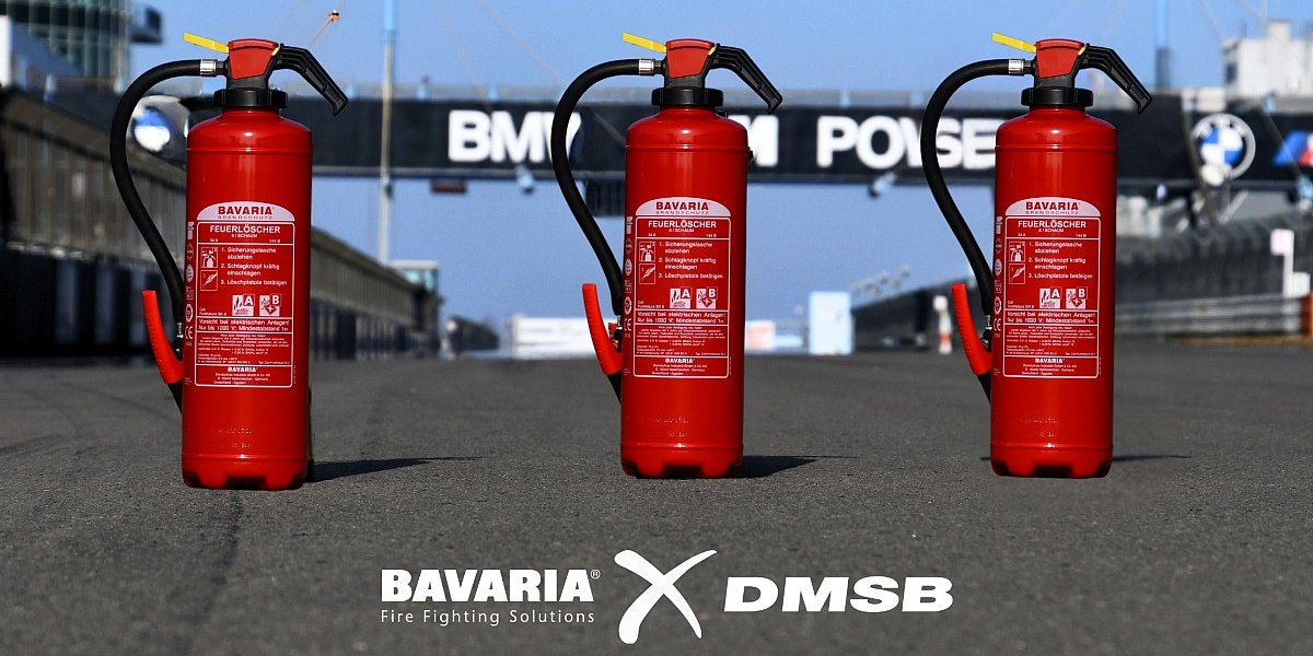 3 rote BAVARIA Fire Fighting Solutions Feuerlöscher stehen auf einer asphaltierten Rennstrecke bei blauem Himmel. Unten die Logos von BAVARIA x DMSB in weiß.