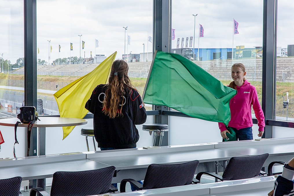 Discover Motorsport – Young Women's Edition 2026 in Oschersleben Zwei Teilnehmerinnen schwenken im Pressezentrum große grüne und gelbe Flaggen und üben so den Einsatz im Ehrenamt.