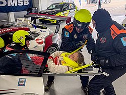 DMSB Medical Days 2026 DMSB Medical Days 2026: Team in dunkelblauen Overalls und gelben Helmen bei Fahrerbergung an weißem Rennwagen; rotes Porsche-Heck sichtbar.