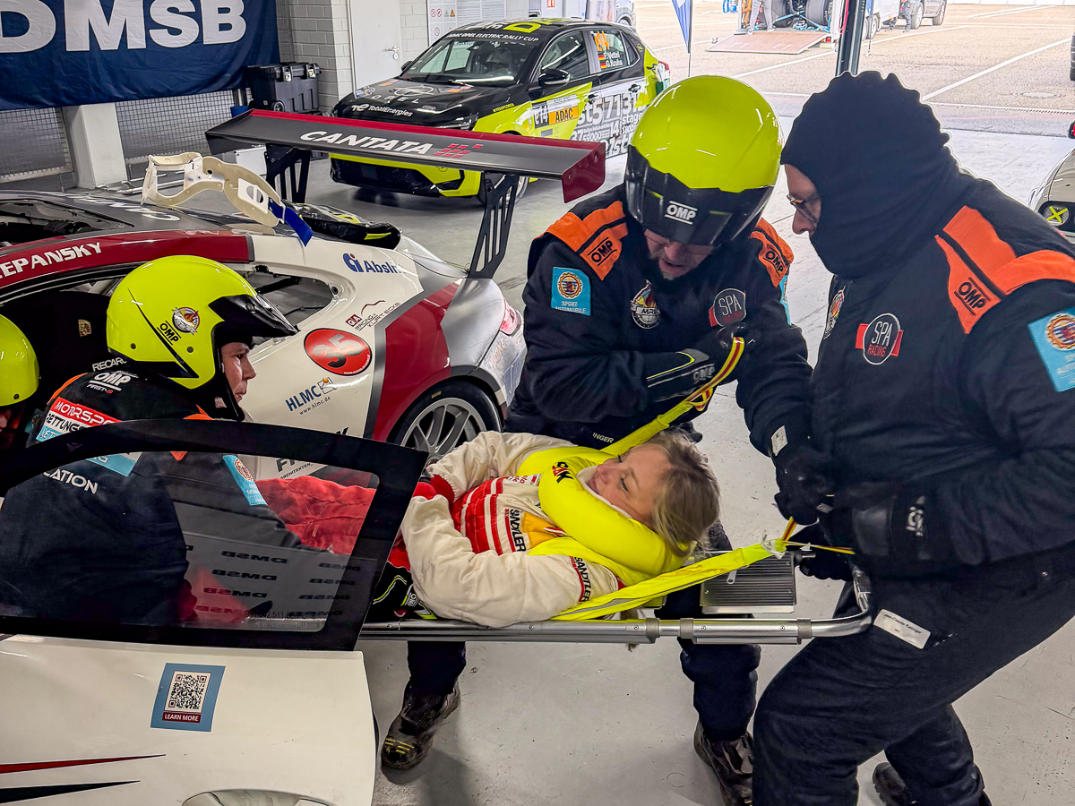 DMSB Medical Days 2026: Team in dunkelblauen Overalls und gelben Helmen bei Fahrerbergung an weißem Rennwagen; rotes Porsche-Heck sichtbar.