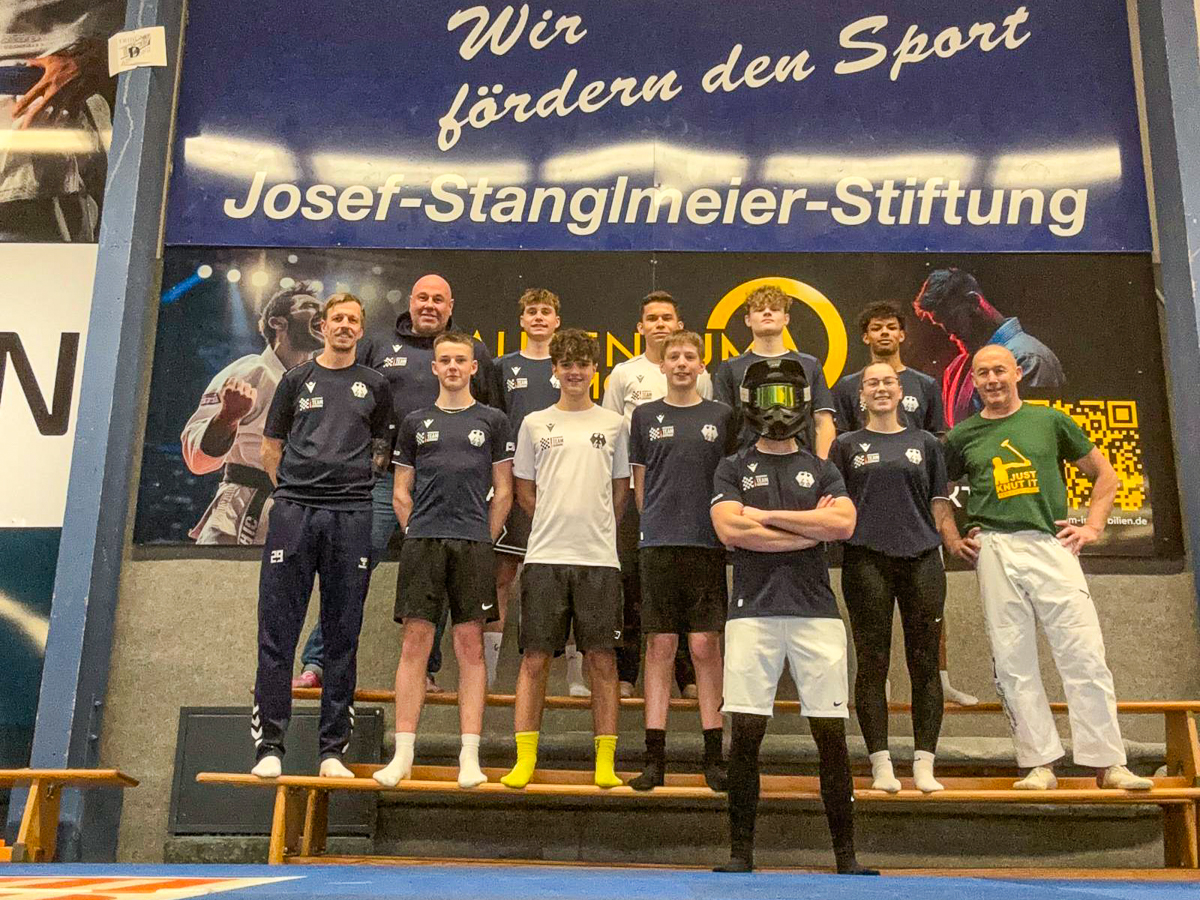 Gruppenfoto des Motorsport Team Germany Speedway beim Saisonauftakt-Fitnesscamp 2026 in Abensberg; 9 junge Fahrer und 3 Betreuer in Teamkleidung in Turnhalle mit Gymnastikboden stehen auf einer Bank nebeneinander aufgereiht vor Josef-Stanglmeier-Stiftung-Banner.