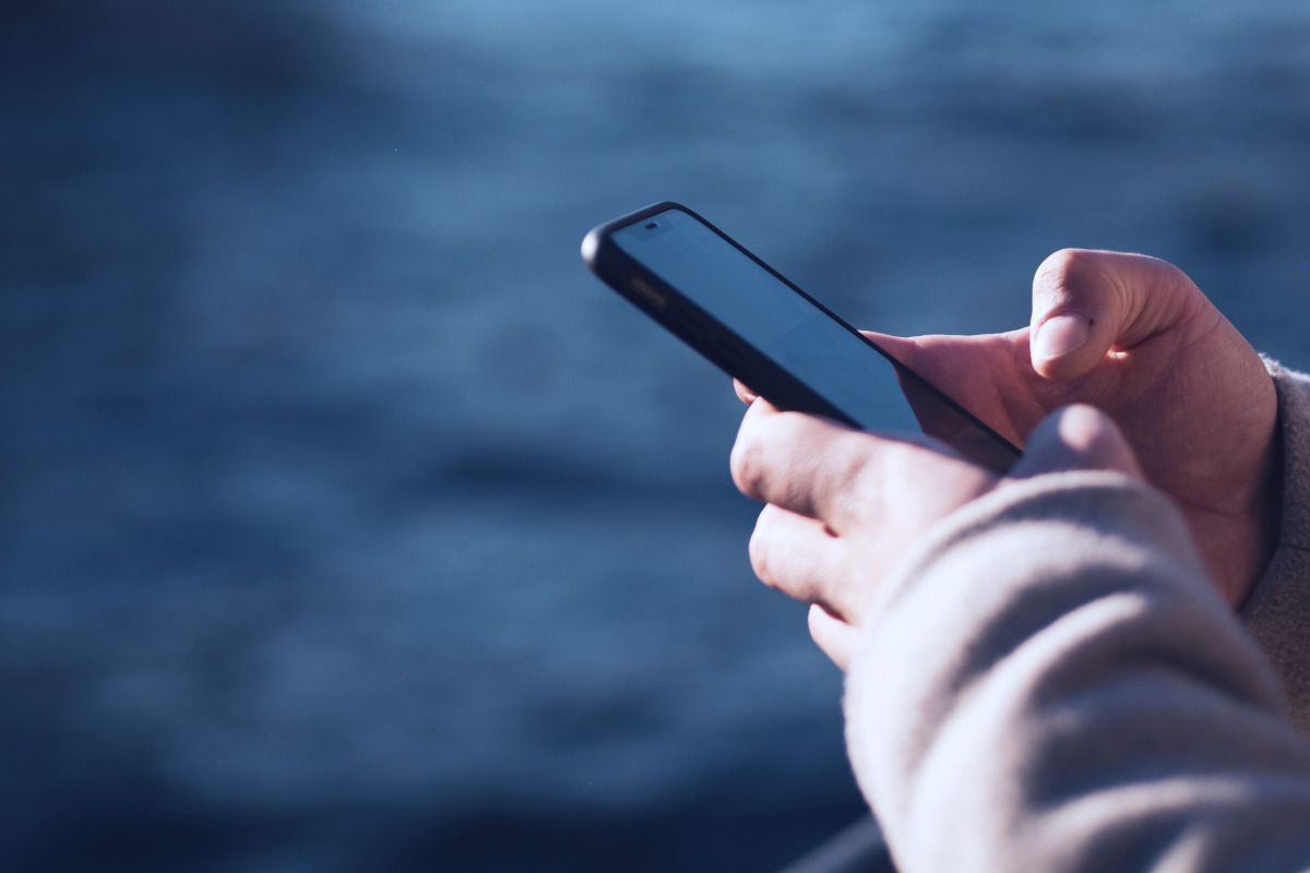 Foto einer Hand, die ein Smartphone hält und auf das beleuchtete Display tippt. Dunkle Dämmerungsstimmung mit blauem Himmel, unscharfem Hintergrund.