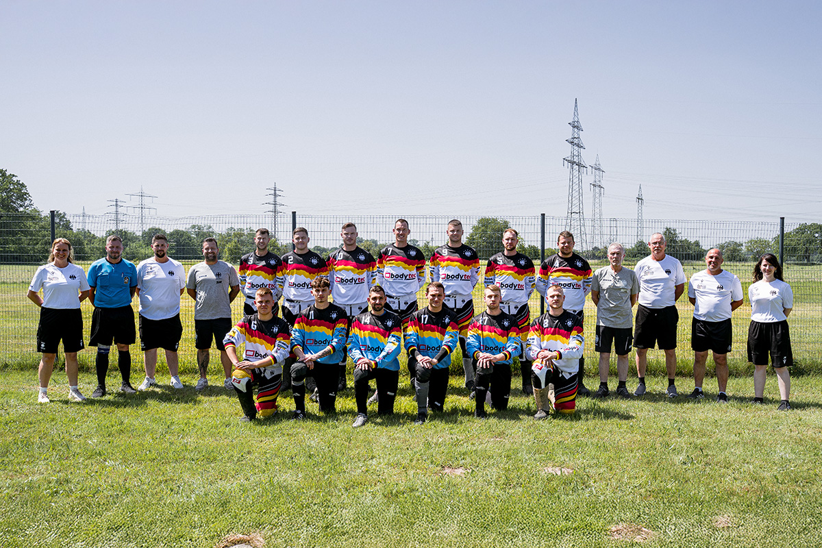 Aufreihung des Motorsport Team Germany Motoball, 2 Reihen Personen mit deutschen Motoball Trickots stehen bzw. knien auf einer grünen Wiese unter freiem Himmel und schauen in die Kamera