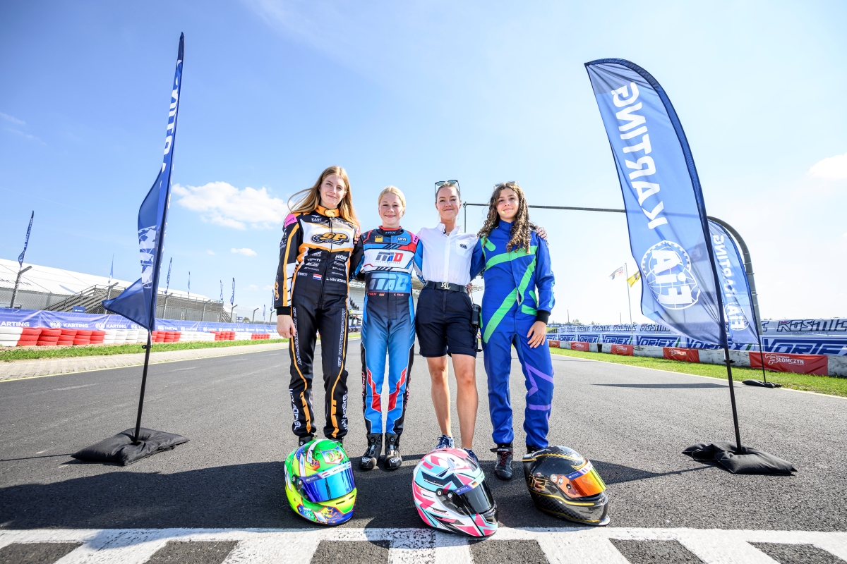 Charlotte Tille bei der FIA Karting Arrive & Drive European Championship 3 Fahrerinnen in Rennanzügen und eine Frau der FIA auf der Rennstrecke, vor ihnen liegen ihre Helme, an der Seite Beach Flags zu FIA Karting