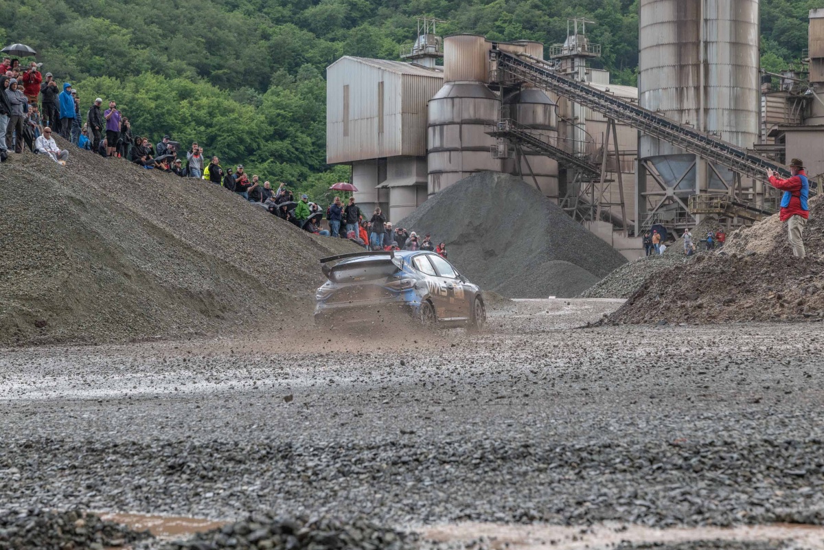 Ein Rallyefahrzeug fährt in einer Schotterkurve auf der Rennstrecke bei der Hunsrück Rallye 2024. Am Rand Zuschauer und ein Fotograf.