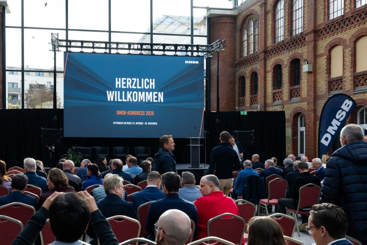 Podium mit Rednerpult und Leinwand, davor Menschen in Sitzreihen, im Hintergrund Glasscheiben mit Sicht nach draußen. Auf der Leinwand steht: Herzlich Willkommen. DMSB-Kongress 2025. Offenbach am Main | 31. Oktober - 02. November 2025.