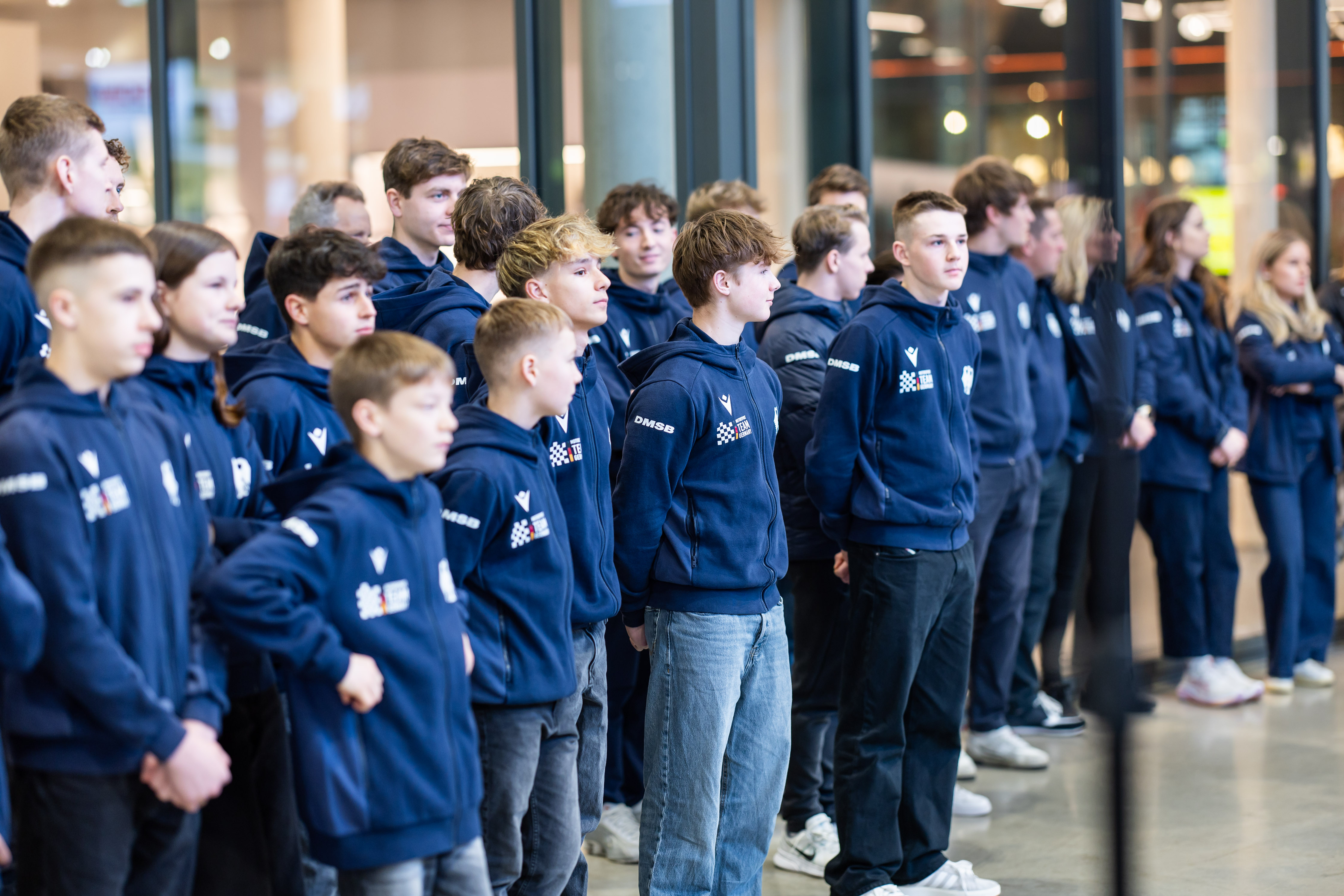 Eine Gruppe von Motorsportlern in einheitlichen dunkelblauen Kapuzenpullis steht drinnen in einer Reihe und blickt in dieselbe Richtung.